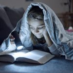 A young girl reading a book under a blanket using a flashlight at night.